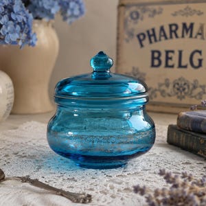 Peut inclure: Un pot en verre turquoise avec un couvercle, orné d'un motif argenté décoratif à la base. Le pot est posé sur un tissu en dentelle blanche, avec une cuillère en argent au premier plan. L'arrière-plan comprend des vases avec des fleurs bleues et une enseigne vintage.