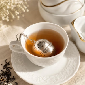 May include: A silver metal spherical tea infuser with small circular perforations rests inside a white ceramic teacup filled with amber-colored tea. The teacup sits on a matching white saucer with a textured rim, accompanied by a silver chain.