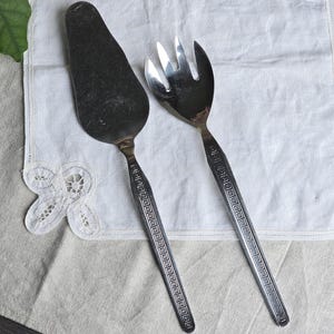 May include: A silver cake server and salad fork set. The server has a wide, flat blade, while the fork has three tines. Both utensils have long handles with a geometric design. The set is displayed on a white linen cloth.