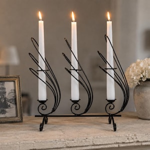 May include: A black metal candelabra featuring three vertical holders for taper candles. Each holder is flanked by curved, wire-like decorative elements that sweep upward. Three white taper candles are inserted and lit, emitting a soft glow.