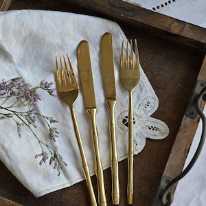 May include: Four gold-coloured forks and knives with bamboo-style handles are arranged on a white cloth. The utensils are placed on a wooden tray with a handle. A sprig of purple flowers is on the left.