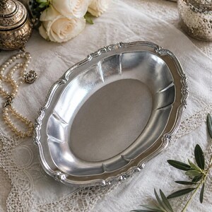 Può includere: Un vassoio ovale argentato con un bordo decorativo smerlato. Il vassoio è esposto su una superficie bianca strutturata, con una collana di perle, un piccolo barattolo decorativo e rose bianche sullo sfondo. Un rametto di foglie verdi è in primo piano.