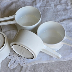May include: Several white enamel measuring cups with long handles are arranged on a linen cloth. The cups have a black rim and a classic design. The image has a vintage aesthetic, reminiscent of a farmhouse kitchen.