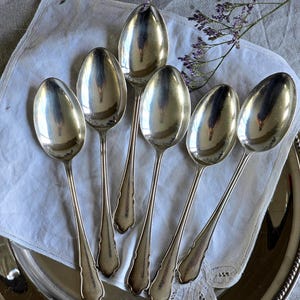 Peut inclure: Six cuillères en argent disposées sur un plateau argenté avec un tissu blanc. Les cuillères ont des cuillères allongées et des manches décoratifs. Les cuillères sont réfléchissantes, et le plateau a un bord perlé.