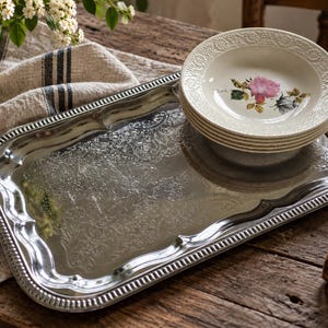 Peut inclure: Un plateau de service rectangulaire en métal argenté avec une bordure gaufrée ornée et une gravure florale centrale, surmonté d'une pile de quatre assiettes en céramique crème avec un motif de rose rose et un bord texturé.