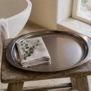 Peut inclure: Un grand plateau de service ovale en métal argenté poli. Sur le plateau repose une serviette en lin beige clair pliée, surmontée d'une branche de feuilles d'eucalyptus vertes, formant un ensemble simple sur un tabouret en bois rustique.