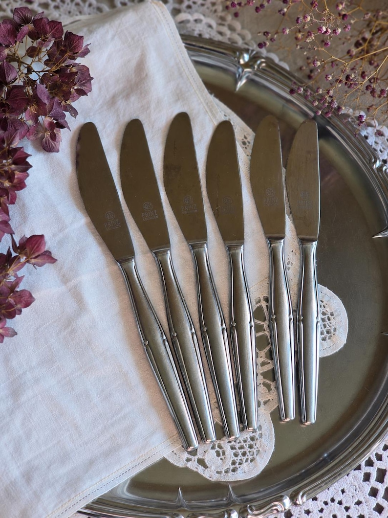 Pode incluir: Seis facas de a&ccedil;o inoxid&aacute;vel dispostas sobre um pano branco e uma bandeja de servir prateada. As facas t&ecirc;m um design simples com um cabo ligeiramente curvo. As l&acirc;minas s&atilde;o lisas e brilhantes, refletindo a luz.
