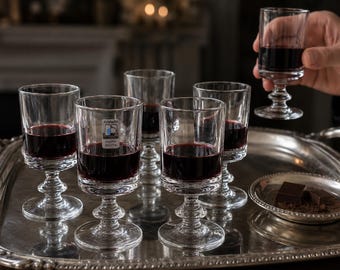 May include: A set of clear glass goblets with short, tiered stems, each filled with dark red wine. One glass is held up, while others sit on a silver tray alongside a small plate containing two dark chocolate squares and cocoa powder.