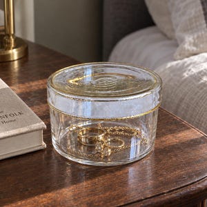 May include: A clear glass trinket box with a circular lid featuring a gold-toned rim and textured detailing. Inside the container, a gold-colored metal ring and a delicate gold chain necklace are visible, resting on the bottom of the glass vessel.