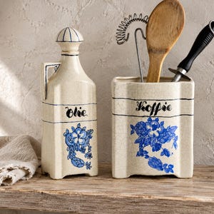 Pode incluir: Uma garrafa de azeite em cerâmica cor de creme com tampa e um suporte quadrado para utensílios a condizer. Ambas as peças apresentam um padrão floral azul, linhas horizontais pretas e as palavras Olie e Koffie escritas a preto na frente.