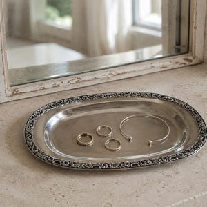Könnte beinhalten: Ein ovales, silberfarbenes Metalltablett mit einem verzierten, geprägten Rand, der komplizierte Schnörkelmuster aufweist. Auf der glatten, reflektierenden Oberfläche des Tabletts liegen drei Schmuckstücke: zwei schlichte, dünne Ringe und ein offener Armreif.