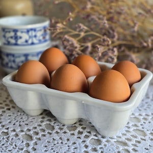 May include: Six brown eggs in a white ceramic egg carton, set on a white lace tablecloth. A stack of blue and white patterned mugs and dried flowers are visible in the background.
