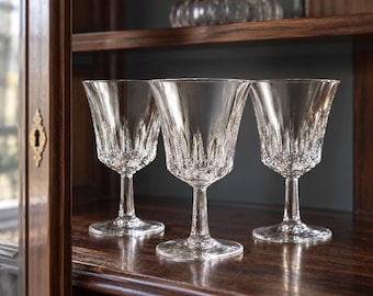 May include: Three clear glass goblets featuring a vertical cut pattern along the lower bowl and a smooth, flared rim. Each glass stands on a slender stem with a circular base, showcasing a reflective, faceted design that catches the light.