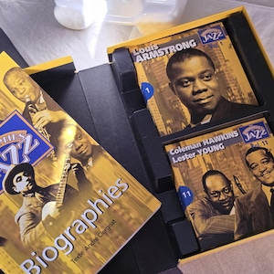 May include: A yellow box set featuring jazz biographies and CDs. The box includes CDs of Louis Armstrong and Coleman Hawkins with Lester Young. The box set is titled "Les Triomphes du Jazz" and "Biographies".