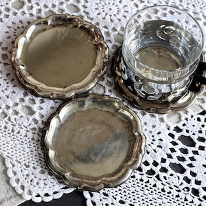 May include: Three silver-toned coasters with scalloped edges. One coaster holds a clear glass with water. The coasters are on a white lace tablecloth. The coasters have a vintage, antique appearance.
