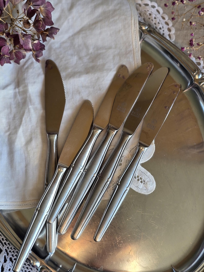 Pode incluir: Um conjunto de facas de manteiga de a&ccedil;o inoxid&aacute;vel com borda serrilhada, dispostas em uma bandeja de servir prateada. As facas t&ecirc;m um design elegante e alongado e s&atilde;o colocadas em um pano branco. Flores roxas secas ao fundo.
