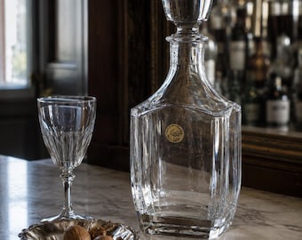 May include: A clear glass decanter with a matching stopper, a crystal wine glass with a faceted stem and bowl, and a small silver-toned footed bowl containing three whole walnuts. The decanter features a circular gold-colored label on its front.