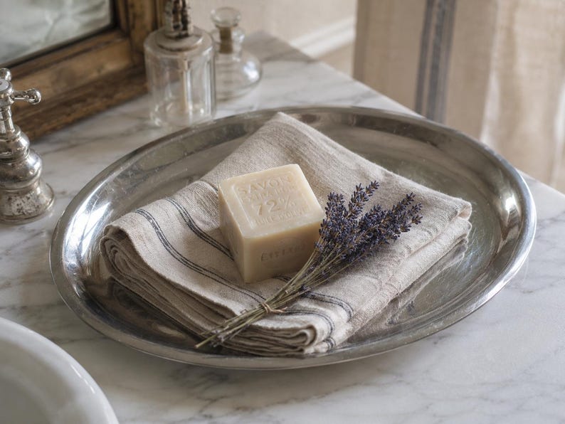 May include: A polished silver-toned oval metal tray holding a folded beige linen cloth with thin dark stripes. A square block of beige soap embossed with the text SAVON DE MARSEILLE 72% HUILE D'OLIVE sits on the cloth next to a bundle of dried lavender.