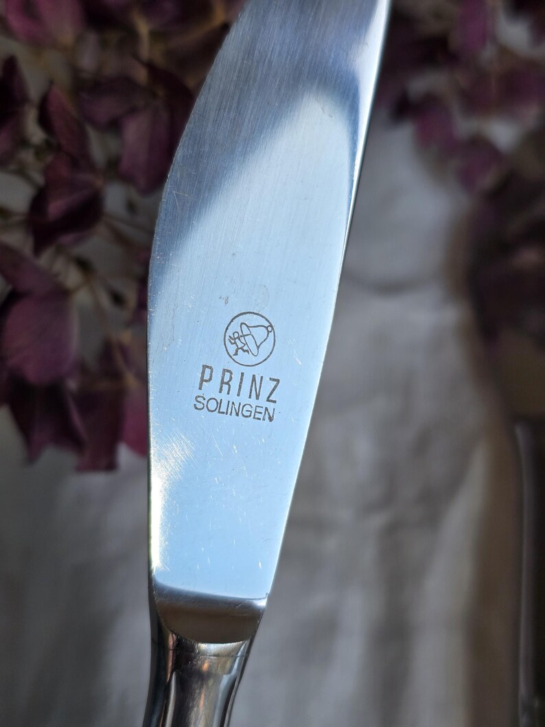 Pode incluir: Close-up de uma faca de manteiga de a&ccedil;o inoxid&aacute;vel com uma l&acirc;mina arredondada. A faca tem a inscri&ccedil;&atilde;o "PRINZ SOLINGEN" e um pequeno emblema. O fundo est&aacute; desfocado, com toques de flores roxas.