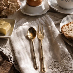 Può includere: Una scena della colazione con un uovo alla coque in un portauovo bianco, una noce di burro e una tazza di tè. Un cucchiaio e una forchetta dorati sono appoggiati su un tovagliolo di lino bianco con bordi in pizzo.