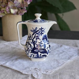 Può includere: Una brocca in ceramica bianca e blu con coperchio. La brocca presenta un motivo floreale blu. Il manico e il beccuccio sono bianchi. È appoggiata su un panno bianco bordato di pizzo.