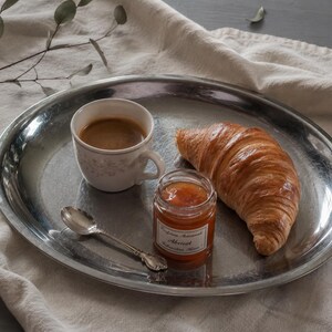 May include: A round, polished silver-toned metal tray holding a white ceramic cup filled with coffee, a golden-brown croissant, a small glass jar of apricot jam with a white label, and a small silver-toned metal spoon resting on the tray surface.