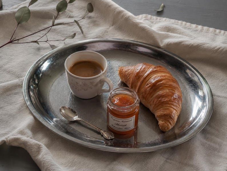 May include: A round, polished silver-toned metal tray holding a white ceramic cup filled with coffee, a golden-brown croissant, a small glass jar of apricot jam with a white label, and a small silver-toned metal spoon resting on the tray surface.