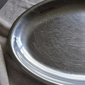 May include: An oval-shaped silver-toned metal serving platter with a smooth, reflective surface. The platter features a wide, slightly raised rim and shows visible fine surface scratches across the center, indicating previous use and wear.