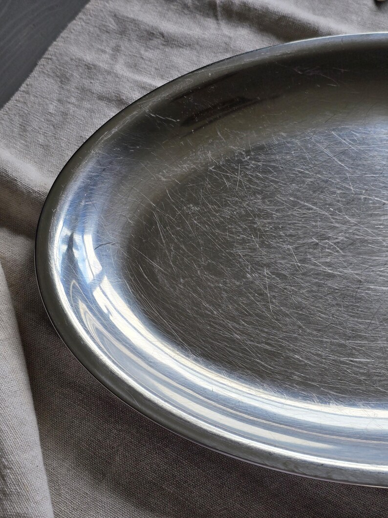 May include: An oval-shaped silver-toned metal serving platter with a smooth, reflective surface. The platter features a wide, slightly raised rim and shows visible fine surface scratches across the center, indicating previous use and wear.