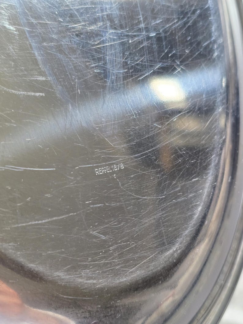 May include: A circular, polished silver-toned metal tray with a reflective surface showing numerous fine scratches. The center of the tray features the stamped text REPPEL 18/8, indicating the material composition of the metallic kitchenware item.