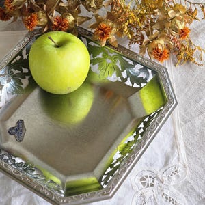 Könnte beinhalten: Ein silberner, achteckiger Tablett mit verzierten, floral gemusterten Kanten hält einen grünen Apfel. Getrocknete orange und gelbe Blumen sind über dem Tablett angeordnet. Das Tablett steht auf einem weißen, spitzenbesetzten Tuch.