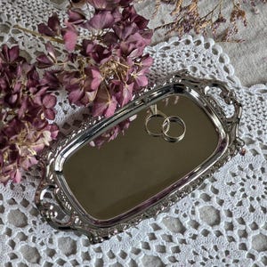 May include: A silver rectangular tray with decorative edges, holding two wedding rings. The tray is set on a white lace doily, with dried purple flowers in the background. The rings are gold in colour.