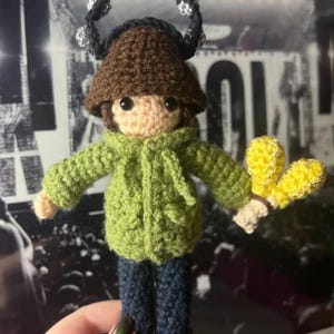 Könnte beinhalten: Handgefertigte Häkelpuppe mit braunem Hut, grüner Jacke und blauer Jeans. Die Puppe hat einen schwarzen Heiligenschein und hält gelbe Blumen. Der Text "LIAM GALLAGHER CROCHET PATTERN" ist oben zu sehen.
