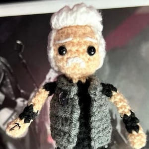 May include: Crocheted doll of a man with white hair and mustache, wearing a gray vest and black pants. The doll has black eyes and is holding a pose. The doll's pants are decorated with silver studs. The text "@Rock.N.Sash.Art" is visible.