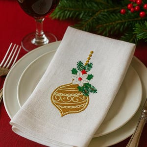 Può includere: Tovagliolo di lino bianco con un ricamo dorato a forma di ornamento natalizio, con un fiore bianco, foglie verdi e bacche rosse. Il tovagliolo è piegato su un piatto bianco, con un bicchiere di vino e decorazioni festive sullo sfondo.