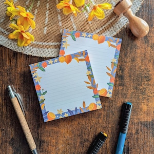 Peut inclure: Deux blocs-notes lignés bleus et blancs avec une bordure de fruits d'agrumes et de fleurs. Les blocs-notes sont sur une surface en bois avec une fleur jaune, un tampon en bois et deux stylos.