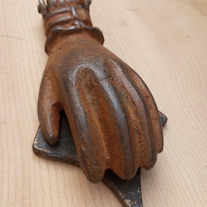 Puede incluir: Un aldabón de hierro oxidado con forma de mano humana. La mano está colocada con los dedos apuntando hacia abajo, descansando sobre una placa de impacto metálica triangular. La superficie del metal muestra una pátina marrón oxidada en todo.