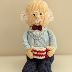 May include: A knitted doll depicting an elderly gentleman with glasses, a bow tie, and a light blue sweater. The doll holds a red and white striped mug. The doll wears navy blue trousers and maroon and green patterned slippers.