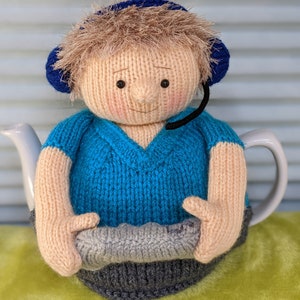 May include: A knitted tea cosy in the shape of a person wearing a blue headset and a blue V-neck sweater. The cosy has light brown hair and a neutral skin tone. The person's hands are holding a grey knitted object.
