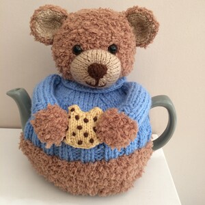 May include: A knitted teddy bear teapot cosy. The cosy is brown and tan with a blue sweater and a cookie applique. The teapot cosy is designed to fit a standard teapot and keep tea warm.