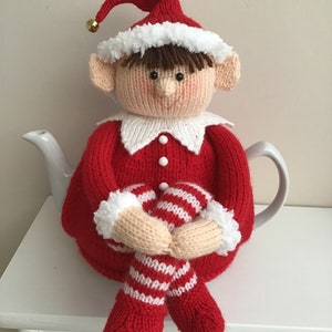 May include: A knitted Christmas elf tea cosy. The cosy is red with white trim and features an elf face with brown hair, beige ears, and a red hat with a gold bell. The elf is holding striped legs.