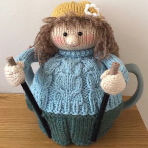 May include: A knitted teapot cosy in the shape of a doll. The doll has brown curly hair, a yellow hat with a white flower, and a light blue sweater. The cosy has black pipe cleaner arms holding beige knitted ski poles.