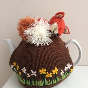 May include: A brown knitted tea cosy with a sleeping fox design. The cosy features a pom-pom tail and a floral embroidered border with yellow and white flowers. The fox is orange and white.