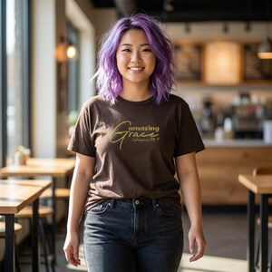 Puede incluir: Una mujer con una camiseta marrón con las palabras "amazing Grace" en letras doradas. El texto "Ephesians 2:8-9" está debajo. Lleva vaqueros azul oscuro y tiene el pelo morado.