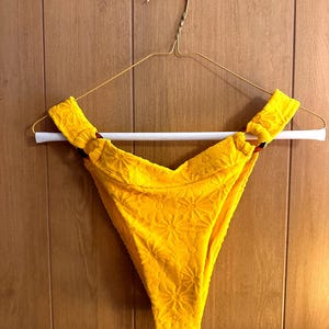 May include: Yellow bikini bottom with a floral pattern and two gold rings on the sides.