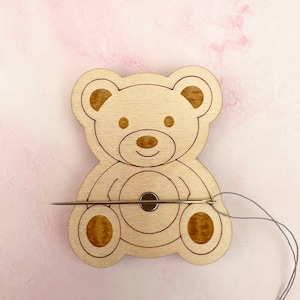 May include: Wooden bear-shaped thread holder with a needle and thread going through it. The bear has a brown nose and brown eyes.