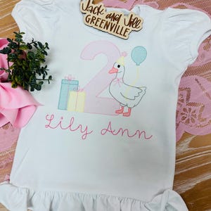 May include: White short-sleeved shirt with a ruffle hem. The shirt features an embroidered design of a goose holding a balloon, the number 2, and the name "Lily Ann". The shirt also has the text "Jack and Jill Greenville".