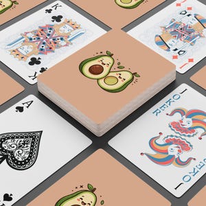 May include: A deck of playing cards featuring a tan card back with a cartoon avocado illustration. The surrounding cards display traditional face card designs, a black spade ace, and a colorful joker card with blue, yellow, and red patterns.