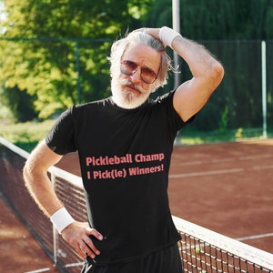 Könnte beinhalten: Ein Mann trägt ein schwarzes T-Shirt mit dem Text "Pickleball Champ I Pick(le) Winners!" in rosa Schrift. Er steht auf einem Pickleball-Platz.