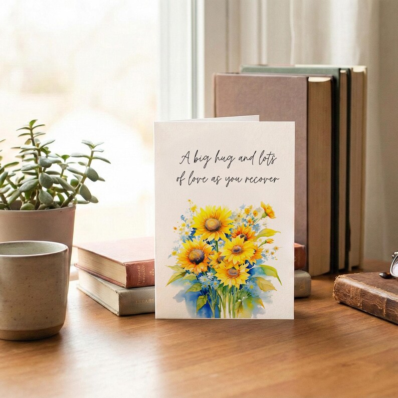 A greeting card with a watercolor illustration of sunflowers sits on a wooden desk, accompanied by a mug, a stack of books, and a potted plant.